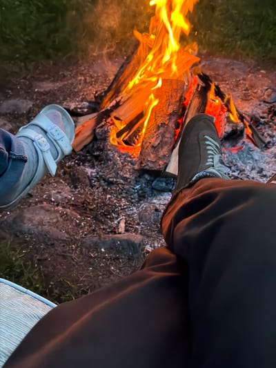 This is a first-person perspective photo of two people sitting by a campfire in Saulkrasti, Latvia. The campfire is in the center of the frame, with bright orange flames and glowing embers. The ground around the campfire is covered in dirt, ash, and rocks. To the left, a person is wearing a light-colored sandal and blue jeans. To the right, another person is wearing dark sneakers and dark pants. The background shows some grass and vegetation. The overall scene depicts a relaxed evening gathering around a fire.