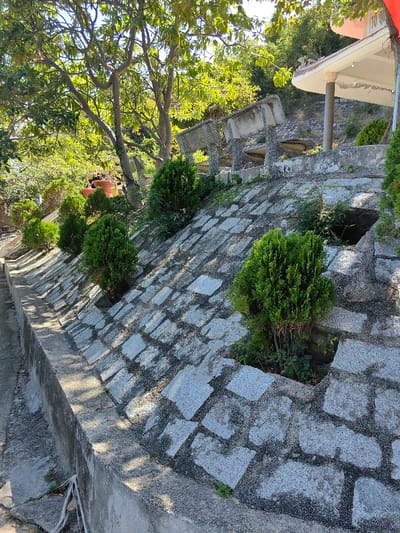 A terraced stone wall climbs a gentle slope, punctuated by squat, verdant shrubs sprouting from square, rocky beds. A concrete retaining wall and narrow trough run along the lower edge. Above, concrete picnic tables and a pavilion with a partially visible flag are nestled amidst trees and greenery. The scene is bathed in sunlight, creating distinct shadows.