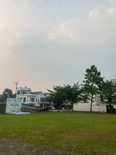 The image depicts a street scene in Cibinong, Indonesia, near the Sentul City area, characterized by modern white houses with blue accents and a large expanse of manicured green lawn in the foreground. A large banner that reads "Selamat Hari Raya Idul Adha" (Happy Eid al-Adha) is prominently displayed on the left side of the image, accompanied by decorative elements resembling crescent moons and stars, and the date "10 Dzulhijjah 1443 H". Several trees, some tall and slender, others with fuller canopies, are interspersed between the houses and along the property lines. A street light stands to the left of the houses, and a paved walkway is visible at the bottom of the frame. The sky is overcast, suggesting a hazy or cloudy day. A few cars, including a white van and a dark-colored SUV, are parked in the background, near the houses. The overall impression is of a residential development or a community area, possibly decorated for a religious holiday.