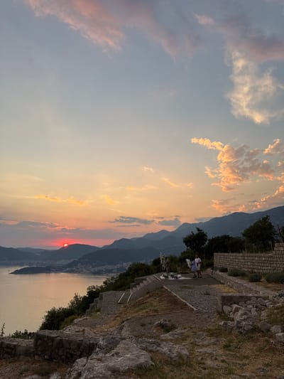 This is a vertical, eye-level, medium shot captured at dusk from a high vantage point overlooking a bay and distant mountains. The sun, a bright red orb, is setting behind a mountain range, casting a warm, orange glow across the sky and reflecting on the water. Scattered clouds are illuminated with pink and orange hues.

In the foreground, a stone terrace overlooks the bay. A paved path leads to a gathering of people. Two individuals, a man and a woman, stand together near a decorative floral arrangement. The man is wearing a white shirt and the woman is in a blue dress. Another woman, standing further away, appears to be observing them. A low stone wall runs along the edge of the terrace, and scattered rocks and dry vegetation are visible.

The midground features a city nestled along the coastline, illuminated by the fading light. Trees and the rugged terrain of the mountains form a backdrop. The distant mountains are silhouetted against the colorful sky.

The image appears to be taken in Čelobrdo, Montenegro, likely from a viewpoint offering panoramic views of the Budva Riviera. The overall mood is serene and picturesque, capturing the beauty of a sunset over the Adriatic Sea.