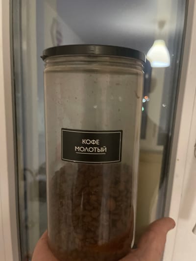 A clear plastic jar filled with coffee beans sits horizontally in front of a window. The jar has a black lid and a black label with white text. The text on the label reads "КОФЕ МОЛОТЫЙ", which translates to "GROUND COFFEE" in Russian. Outside the window, a blurry night scene is visible with lights reflecting. A hand, likely belonging to a person, is holding the jar from the right side of the image. The scene appears to be indoors, possibly in a kitchen or on a windowsill. The lighting suggests it is nighttime. The overall mood is still and domestic.