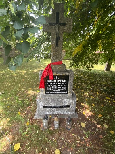 This is a close-up, eye-level shot of a tombstone in the village of Puchły, Poland. The tombstone is made of grey granite with a black plaque in the center. The plaque has text in Cyrillic script, indicating a burial of individuals with the surname "Димитруки" (Dimityuki). The text lists names such as "Варвара" (Varvara), "Иосиф" (Iosif), "Агафия" (Agafiy) and "Деомит" (Deomit) along with their lifespans in years. Below this, it mentions "Петро Иаков, Евфросиния Макарий" (Petro Iakov, Efrosinia Makariy) and a year "1918 г." (1918). A red ribbon is tied around the upper part of the tombstone, and a crucifix is affixed to the top of the stone. In front of the tombstone, three candle holders, two glass and one metal, are placed on the ground amidst fallen leaves and grass. The background shows trees with green and yellowing leaves, and a partially visible stone wall and another tombstone to the right, suggesting it is a cemetery. The lighting is natural, coming from above and to the right, creating some shadows.