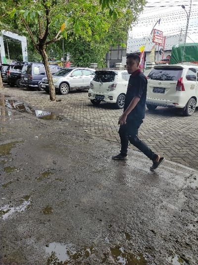 This image captures an outdoor daytime scene in Kecamatan Asembagus, Indonesia, likely an urban or suburban area. The main subject is a young man, possibly of East Asian descent, positioned slightly to the left of the center. He is dressed in a dark short-sleeved collared shirt, dark trousers, and dark shoes. He is captured in a dynamic pose, with his left leg extended straight backward and his right leg bent, giving the impression of him balancing, mid-action, or perhaps playfully attempting a kick or a suspended movement. He is looking towards the camera with a gentle smile.
The setting consists of a paved ground surface on the left, possibly made of bricks or blocks, where several cars are parked. The right side of the ground is a rougher, concrete or asphalt-like texture, heavily covered in mud and standing water, suggesting recent rainfall or poor drainage. In the background, multiple white cars, resembling compact SUVs or minivans, are parked. Further behind them, green trees and foliage are visible, along with parts of buildings and what appears to be a commercial sign with red and white elements, though no specific text is discernible. The overall lighting suggests it is bright daylight. The scene appears to be an everyday moment, perhaps on a street or parking area adjacent to shops or residences, marked by the wet and muddy ground conditions.