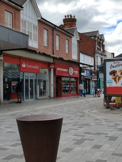 A street scene in Sandwell, United Kingdom, depicts a row of retail establishments along a paved pedestrian walkway. The buildings are constructed primarily of red brick, with some featuring white trim and large bay windows. Shops visible include Santander, Entertainment Exchange, and a mobile phone retailer. The walkway is paved with grey and darker grey rectangular tiles laid in a pattern.
In the foreground, a dark brown, conical planter stands to the left. A man in a dark jacket and shorts is using an ATM outside the Santander bank. Further down the street, two people are pushing a stroller. To the right, a large JCDecaux advertising billboard for "Kinder Bueno" displays images of ice cream cones. Various boxes and debris are scattered on the pavement near the billboard, and some litter is visible throughout the scene. The sky is overcast with clouds.