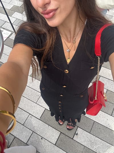 A medium shot captures a woman from the chest down, standing outdoors on a sidewalk paved with light gray and white rectangular tiles. She is wearing a black, ribbed, V-neck dress with short sleeves and gold buttons down the front. The dress has two gold-buttoned pockets on the chest. She is also wearing black sandals with white toenail polish. A gold bracelet is visible on her left wrist, and a bright red handbag with a gold chain strap is slung over her right shoulder. She is also wearing a necklace with multiple gold pendants, one of which is a small, gold-toned letter "Y" with diamonds. Her hair is long and brown. The image provides a clear view of the woman's outfit and accessories against the backdrop of the tiled sidewalk. The location is identified as the city of Aventura, United States of America, though no specific landmarks are visible in the frame to confirm this.