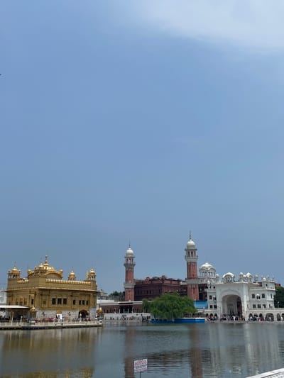 The image depicts the Golden Temple, also known as Sri Harmandir Sahib, located in the city of Amritsar, India. It is a prominent Sikh gurdwara, the holiest of Sikh shrines. The photograph is taken from across the Sarovar, a large pool of water surrounding the temple complex.
In the foreground, the water reflects the sky and the structures. The Golden Temple, a magnificent golden-domed building, is situated on a causeway in the middle of the Sarovar. It is a multi-story structure with intricate architectural details, including domes and minarets, all covered in gold leaf. People are seen walking on the causeway leading to and from the temple.
To the right of the Golden Temple, a large complex of white buildings is visible, featuring arches and domes. Behind this white complex, a tall, red brick minaret rises, topped with a white dome. Further to the right, another building, also part of the complex, is visible. The architecture of these adjacent buildings complements the grandeur of the Golden Temple.
The sky above is a pale blue with some scattered clouds. The overall scene conveys a sense of spiritual serenity and architectural splendor. The image captures a significant religious and cultural landmark of Amritsar.