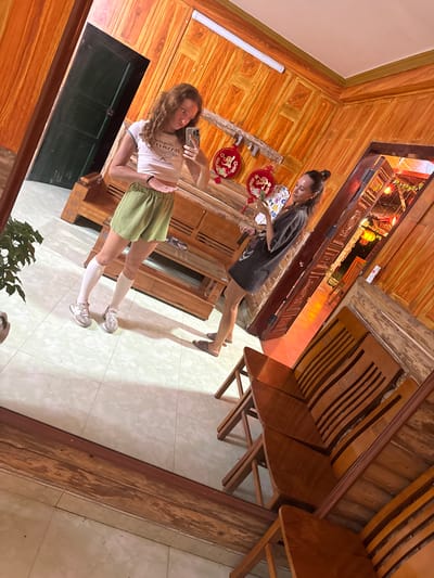 Two women are captured in a mirror selfie within a wooden-structured building. One woman wears a white top and green shorts, while the other is in a dark-colored, knee-length garment. The location is Mèo Vạc Commune, Vietnam. Architectural details include wooden paneling and traditional decorations. The women are in the act of taking a photograph using a smartphone. The background is a mix of traditional Vietnamese decor and furniture. The floor is tiled.