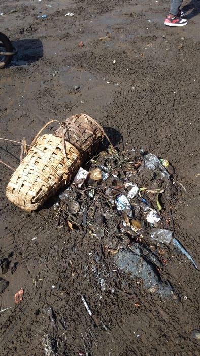 The image shows a large woven basket lying on its side in the dirt. The basket appears to have been used to transport or store items. It is filled with debris, including plastic, glass, and metal objects. The ground is covered in dirt and mud, and there is a footprint in the upper right corner of the image. The image was taken at a location in Limbe, Cameroon, near the coordinates 4.005, 9.21.