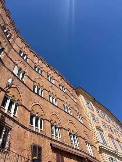 The image shows a low-angle view of the brick facade of a tall building in Siena, Italy. The building is several stories high and features rows of windows with white frames and decorative brickwork surrounding them. The sky is a clear, bright blue. The building's design includes arched elements above each window, creating a pattern along the facade. Some windows have closed shutters. A street light and other architectural details are visible on the right side.