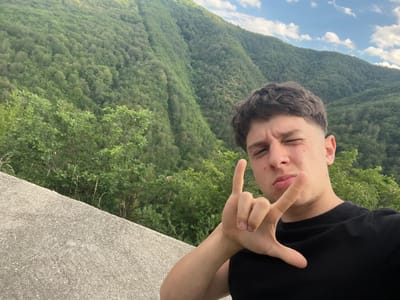 The image is a selfie taken outdoors in a mountainous, forested area. The primary subject is a young man with short, dark, styled hair, wearing a black t-shirt. He is looking towards the camera and winking, with his right hand making a gesture that resembles the "rock on" sign.

The background features a steep, verdant mountainside covered in dense green trees. Above the mountain, a blue sky with scattered white clouds is visible. In the foreground, a rough, textured concrete surface runs diagonally across the bottom left of the frame.

The lighting suggests it is daytime, likely afternoon, given the bright sky and the even illumination of the scene. The overall impression is one of a casual, spontaneous moment captured while enjoying the natural scenery. There are no visible text elements in the image. The location is identified as Pechinska, Bulgaria.