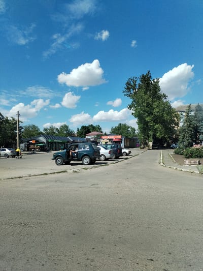 In Nova Odesa, Ukraine, a scene unfolds on a sunny day with a bright blue sky dotted with puffy white clouds. The foreground shows a paved road with some wear and tear. To the left, a silver sedan is parked. Further back, a dark green SUV with its driver's door open is positioned in front of a row of low-rise buildings that appear to be shops or businesses. One of these buildings has a sign that reads "Hub". To the right of the SUV, a white sedan is parked. Beyond these vehicles, the landscape transitions to trees and a multi-story building. A tall, mature tree with dense foliage stands prominently in the middle ground. The area is characterized by a mix of commercial and residential structures, with ample open space and vegetation.