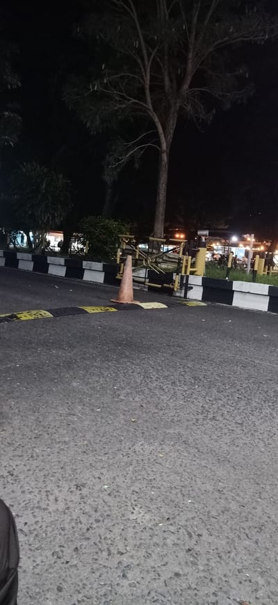 At night in Tambontunong, Indonesia, an orange traffic cone stands on an asphalt road. The road is marked with black and yellow speed bumps. In the background, a metal barrier with yellow and black markings is visible. Beyond the barrier, trees and some buildings with illuminated windows can be seen. The scene is dimly lit, suggesting it is late at night.