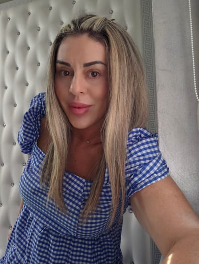 A woman with blonde hair is leaning against a white upholstered headboard. She is wearing a blue and white gingham dress. The headboard has tufted diamond patterns with clear crystals. To the right of the woman is a textured grey wall with a Greek key pattern border. There are no other people or objects in the frame, and the environment suggests an indoor setting, possibly a bedroom. The image was taken in the city of Carpi, Italy.