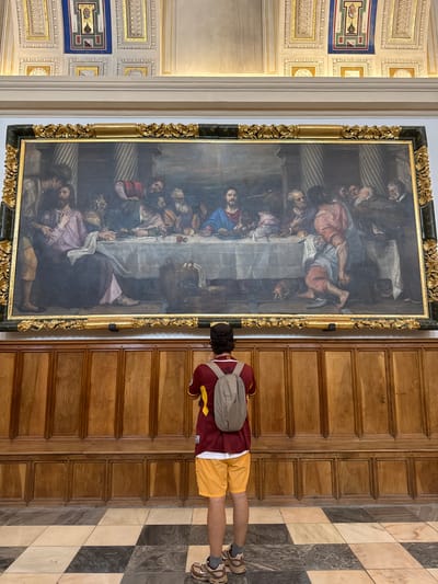 In the Hall of the Kings within the Royal Site of San Lorenzo de El Escorial, Spain, a person stands before a large oil painting depicting "The Last Supper." The painting is framed by an ornate gold frame and hangs on a wall adorned with wooden paneling and benches below. The floor is a checkerboard pattern of light and dark stone tiles. The person is wearing a maroon and yellow shirt, yellow shorts, a grey backpack, and sneakers. They are facing away from the camera, looking intently at the artwork, which features Jesus and his disciples gathered around a table laden with food. The scene is illuminated by natural light, casting soft shadows. The ceiling above the painting is decorated with intricate stucco work.