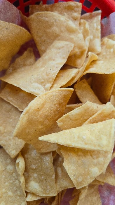 A close-up shot displays a pile of golden-brown tortilla chips, likely made from corn, filling a red plastic basket. The chips are mostly triangular, varying in size and shape. Some chips are stacked on top of others, creating depth and texture. The chips exhibit a slightly rough surface. The warm tones of the chips contrast with the basket's red hue.