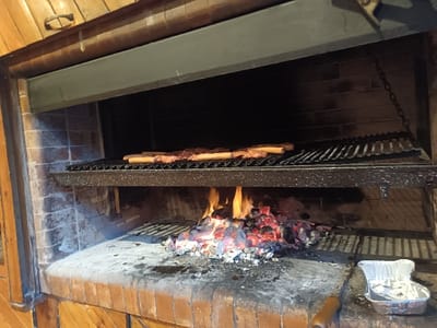 A brick fireplace holds a fiery charcoal bed below a metal grate. Sizzling meat, possibly burgers, rests atop the grate, cooking over the intense heat. The stone and brick interior provides a rustic setting, while an aluminum foil pan sits nearby on the hearth, suggesting preparations are underway. The scene evokes a sense of traditional cooking and warmth.