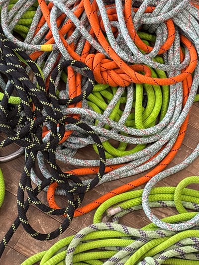 A substantial, disorganized pile of coiled ropes is depicted resting on a brown wooden plank surface. The ropes exhibit various colors and patterns, including bright orange with dark flecks, neon green (some solid, others with dark stripes), greyish-white with red and blue flecks, and black with bright yellow checkered patterns. One rope segment displays yellow tape, and another light green rope shows a slightly frayed end. The wooden surface beneath the ropes features visible grain and plank seams. No individuals or active interactions are visible. This scene is observed in the city of Tayshet, Russia.