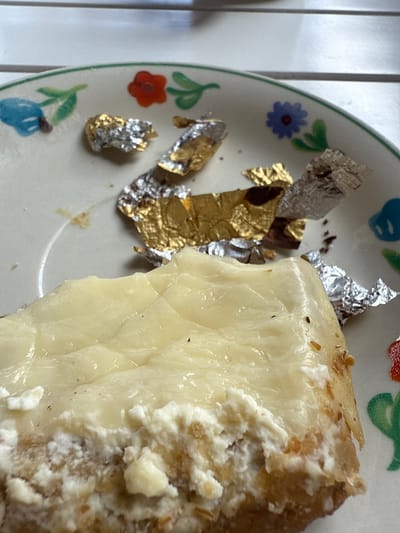 This is a close-up, eye-level, indoor shot of a slice of cheesecake on a white ceramic plate with a green rim and a hand-painted floral pattern. The cheesecake has a light yellow, smooth top layer and a craggy, white, creamy texture underneath. The crust appears to be a light brown color. Scattered on the plate around the cheesecake are crumpled pieces of gold and silver foil, some with dark chocolate residue. The background is blurred, showing horizontal lines which suggest a table or surface. The image does not contain information about Bischofswiesen, Germany, or any specific actions or people.