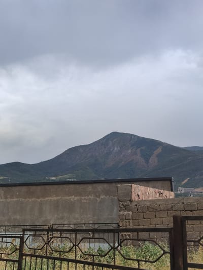 The image depicts a mountainous landscape under a cloudy sky, seen from what appears to be a residential area. In the foreground, a dark metal fence runs horizontally across the lower portion of the frame, with dried vegetation visible behind it. Behind the fence, a low, weathered concrete wall is prominent, with a section of brickwork on its right side. A more substantial, rougher brick wall with a rough concrete cap is situated behind the concrete wall. In the mid-ground, a series of rolling green hills lead to a larger, darker mountain in the background. The mountain has visible scars and patches of lighter soil or rock on its slopes. A scattering of buildings, appearing to be hotels or resorts, are visible on the far right side of the mountain range. The overall atmosphere is somber due to the overcast conditions. The scene is located in Khujakent, Uzbekistan.