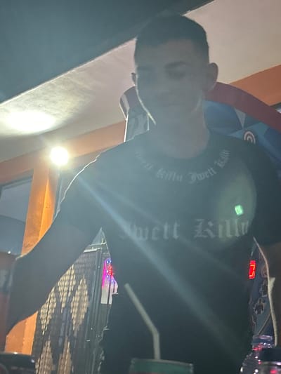 The image shows a young person wearing a dark t-shirt with white lettering on it. The lettering appears to be a repeating phrase with gothic-style font. The person is looking down and to the side, with their face partially obscured by lighting and motion blur. They are standing in what appears to be an indoor or covered outdoor setting, with a bright light source visible in the background. There are other objects visible in the background, including what looks like a decorative wall or screen with a diamond pattern and some illuminated elements. A plastic bottle with a straw is also visible in the foreground. The overall impression is that of a casual setting, possibly a bar or entertainment venue. The image is grainy and appears to have been taken with a mobile phone camera.