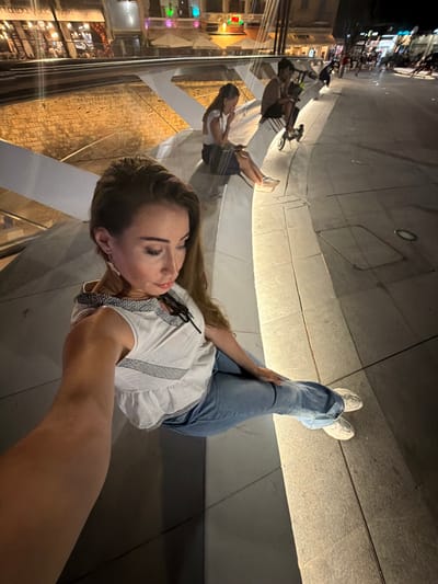 This media file captures a person taking a selfie in an urban environment at night. The person is a woman with long, light brown hair, wearing a white ruffled top and blue jeans. She is positioned in the foreground, with her arm extended to take the photo. Her expression appears relaxed and engaged. In the background, illuminated by artificial lighting, is a street scene with shops and people. The lighting creates a warm glow on the textured, stone-like surfaces in the foreground, which appear to be part of an architectural structure, possibly a walkway or seating area. The overall impression is that of a casual evening outing. The time of day is clearly night, indicated by the dark sky and the artificial lighting. There are no specific weather cues visible. Location cues, such as "Nicosia, Cyprus," are provided, suggesting the setting is an urban area within that region. The reflective surfaces in the image also show a distorted reflection of the scene and the subjects, adding a unique visual element.