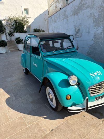 This media file shows a teal-colored vintage Citroën 2CV car parked on a stone-tiled surface. The car is viewed from the front-right, with its hood and passenger side visible. The car has a black convertible top. The front of the car features a chrome grille, a round headlight, and a black bumper with white and black accents. A license plate with the numbers "95522" is visible. The car has "Nº24" written in white on the hood and on the side door.

The setting appears to be outdoors, with a white-painted building in the background. The building has a textured facade and a window. There are also some potted plants and greenery visible. The lighting suggests it is daytime, with strong sunlight casting shadows. The overall mood is bright and sunny. The text on the car reads "Nº24" and "95522". The location context is Monopoli, Italy.