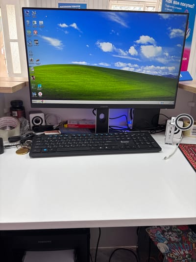 The image depicts a computer desk setup. A large monitor displays the classic "Bliss" wallpaper from Windows XP, showing a green hill under a blue sky with white clouds. On the desk, a black keyboard is positioned in front of the monitor. To the left of the keyboard, there are various items including rolls of tape, a small white speaker, keys with a brass-colored medal, and a dark-colored bottle with a white label. To the right of the keyboard, a USB drive and what appears to be a passport are visible. A mobile phone holder with a ring light attachment is also present on the right side of the desk. In the background, a printer with the "SAMSUNG" logo is visible. The scene is indoors, with a shelf above the desk that has some signage indicating "МИЛЛИОНЫ ТОВАРОВ" (Millions of Goods) and "КАК ВАМ ПОКУПКА?" (How do you like the purchase?). The overall impression is a functional workspace with personal items. There is no indication of the city of Vileyka, Belarus in the image itself.