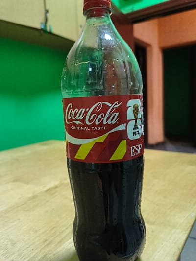 A close-up shot shows a Coca-Cola bottle with condensation on its exterior, placed on a wooden surface. The bottle has a red cap and a label featuring the Coca-Cola logo, the FIFA World Cup trophy, and "ESP" text. The background consists of a blurry green wall and an out-of-focus room with warm-toned walls and doorways. The image appears to be taken indoors, possibly in a domestic setting. No information about the city of Tukums, Latvia is present in the image.