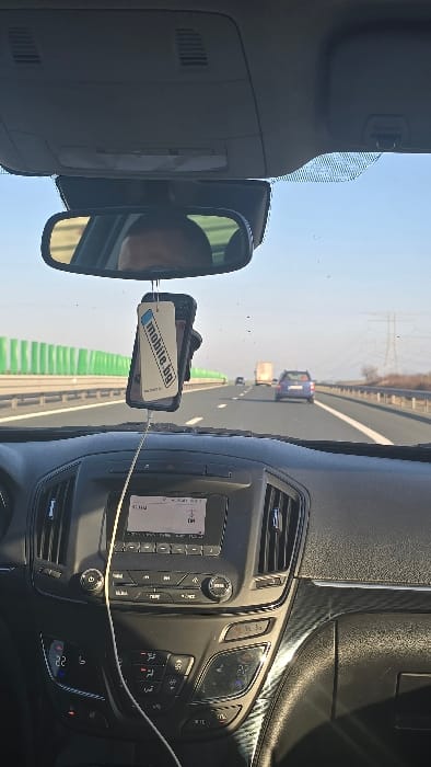 This image depicts a forward-facing view from inside a moving vehicle on a multi-lane highway near Medgidia, Romania, under clear daylight conditions.
**Vehicle Interior:** The foreground features the black dashboard and central console of the vehicle. A white charging cable extends from a smartphone, which is suspended from the rearview mirror. The phone is in a black case, with a beige tag displaying "mobile.bg" in blue text. The vehicle's infotainment system, featuring a screen with text (partially obscured) and various control buttons, is visible below the windshield. Below this, a climate control panel displays "22" degrees on its left side. Vertical air vents flank the infotainment system, and a section of textured trim, possibly carbon-fiber style, is visible on the passenger side of the dashboard. The rearview mirror centrally reflects the upper portion of a human head. The grey overhead console with vents is partially visible in the top of the frame.
**Exterior Environment:** The vehicle is traveling on a paved highway with white lane markings. Ahead, a blue SUV or hatchback is visible in the same lane, and a white truck is further ahead in what appears to be an adjacent left lane. A tall, green sound barrier runs along the left side of the highway. On the right, a metal guardrail separates the highway from a sparse, open landscape. In the distant background on the right, power lines and transmission towers are visible against a clear blue sky.