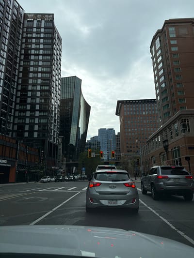 The image shows a street scene in Boston, Massachusetts, USA, on a cloudy day. In the foreground, a silver Hyundai hatchback is stopped at a traffic light. To its right, a silver Jeep Grand Cherokee is also stopped. The road is wet, indicating recent rain.
The background features several tall buildings, characteristic of a downtown urban environment. On the left, a dark-colored skyscraper with many windows dominates the view, with a sign that reads "NOW LEASING". To its right, a modern building with a distinctive curved, glass facade stands out. Further back, more contemporary buildings with reflective glass windows are visible.
Traffic lights are visible, with the red light illuminated, indicating that the vehicles are stopped. Street signs and lane markings are also present on the road. The overall impression is a typical city intersection with traffic and modern architecture.