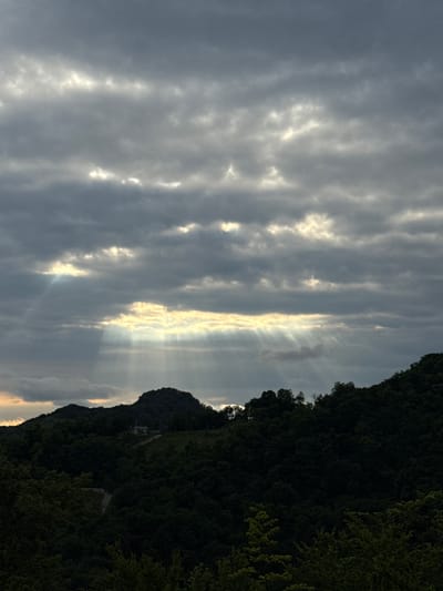 A dramatic vista unfolds, featuring a dense, verdant forest that blankets rolling hills. Above, a turbulent sky is filled with heavy, gray clouds, through which shafts of sunlight pierce, creating a celestial spotlight effect. The light beams illuminate the cloud cover and cast an ethereal glow upon the landscape below. A solitary, light-colored building with a peaked roof is visible on a hilltop, surrounded by the dark, lush foliage. The scene evokes a sense of natural grandeur and a touch of isolation, suggesting a remote and peaceful setting in the Adzhakhy region of Abkhazia.