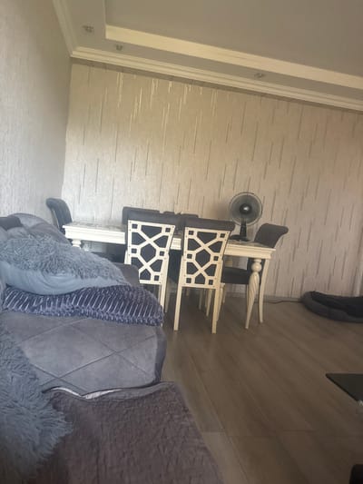 The image shows an indoor dining area with a sofa in the foreground. A dark grey sofa with several pillows is partially visible on the left side of the frame. Behind the sofa, a white dining table with four chairs is set up. The chairs have dark upholstered seats and backrests with a decorative geometric pattern. A black oscillating fan stands on the dining table. The walls are covered with textured, light-colored wallpaper. The floor is made of light brown laminate. In the background, on the right side of the room, a dark pet bed is visible on the floor. The scene appears to be a residential interior.