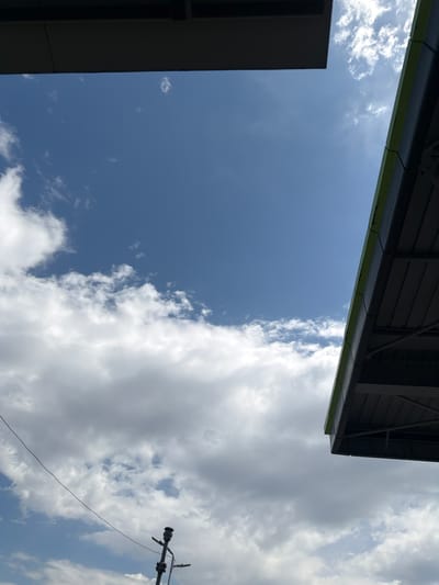 The image shows a view towards the sky from a lower vantage point. Dominating the left side of the frame are large, puffy white clouds partially obscuring a blue sky. A utility pole and wire are visible in the upper left corner. On the right side, a dark gray building edge enters the frame. In the lower portion, a gray structure with a green accent line runs diagonally, suggesting a roof or architectural element. The sky is blue on the right. It is not possible to confirm the specific location of Yoakim Gruevo, Bulgaria from the visual information alone. There are no people in the image.