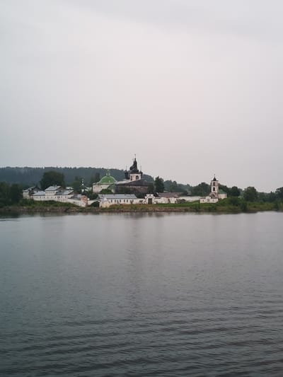 The image captures a wide view of the Goritsy Monastery, located in Goritsy, Russia, from across a body of water. The monastery complex is situated on a gentle slope leading down to the water's edge. Prominent structures include several white buildings with red roofs, a large green-domed church, and a white bell tower with a dark steeple. The architecture appears traditional Russian. The monastery is surrounded by lush green trees and foliage, with a dense forest visible in the background on a distant hill. The sky is overcast and a light gray, contributing to a somewhat muted and serene atmosphere. The foreground is dominated by the dark, rippling surface of the water, reflecting the overcast sky. No people are visible in the image. The overall scene suggests a tranquil, historic religious site.