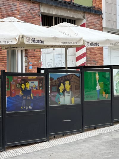 This outdoor scene, captured in Oviedo, Spain, displays a street setting with commercial elements and displayed artworks. On the left side, a large white retractable awning is prominent, providing shade. The awning is adorned with the brand name "Alhambra" in dark blue lettering, and the slogan "CERVEZA HECHA SIN PRISA" (Beer made without hurry) repeated several times. Behind the awning, a building facade constructed of reddish-brown bricks is visible, along with some concrete sections and dark electrical cables running vertically.
To the right, a series of framed artworks are vertically displayed on a black stand, possibly for sale or as decoration. These artworks appear to be personalized caricatures or "Simpsonized" portraits. The topmost visible artwork depicts a character resembling Homer Simpson lying in green grass. Below this, another artwork features a man and a woman, both with yellow skin tones, in what appears to be a bar setting. The word "ЯАВ" (BAR, mirrored) is visible in the background of this piece. The bottom visible artwork similarly shows a man and a woman, also in a bar, with details like a dartboard and a jukebox. The mirrored "ЯАВ" text is present in this artwork as well.
The ground surface, visible on the far right, is a light grey concrete sidewalk with a textured, possibly gridded, pattern. The lighting suggests it is daytime, under clear weather conditions. The overall impression is that of a bustling urban area, possibly near a cafe or bar.