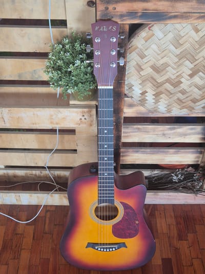The image shows a brown-and-yellow acoustic guitar lying horizontally on a wooden pallet. At the head of the guitar, there is a small potted plant. Behind the head is a woven plate. To the right of the guitar's body is a vase with dried flowers. The background behind the pallet and guitar is a wooden floor. The location is likely the city of Pasay, Philippines.