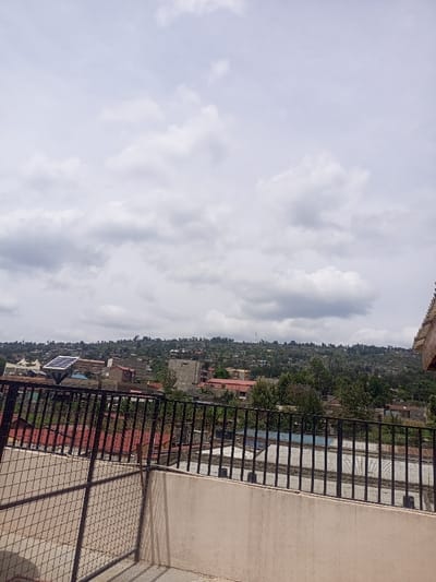 The image displays an elevated panoramic view of the Kiamaina ward in Kenya under an overcast sky filled with white and gray cumulus clouds. In the immediate foreground, a beige concrete parapet wall is visible, topped by a black metal railing with vertical bars extending horizontally across the frame. To the left, a section of a black metal grid fence is also present, angled towards the viewer.

The midground reveals a densely populated urban area characterized by numerous buildings of varying sizes and construction. Many structures exhibit red or corrugated metal roofs. A notable building on the left side of the midground features a prominent blue solar panel array installed on its roof. Towards the right-center, a multi-story building under construction is visible, characterized by its exposed concrete structure and gray facade. Patches of green trees and dense foliage are interspersed among the buildings and on the slopes.

The background consists of undulating hills covered extensively with a mix of buildings and lush green vegetation, indicating further urban or semi-urban sprawl extending into the distance. No individuals, discernible actions, or direct interactions are visible within the scene.