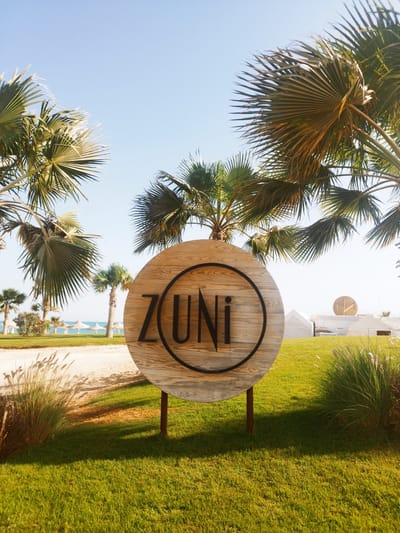This media file captures a bright, sunny daytime scene of a tropical resort area, specifically in Ras Abu Soma, Egypt.
The central subject is a large, circular wooden sign placed prominently in the foreground. The sign features the word "ZUNI" in bold, black, sans-serif letters, encircled by a thin black ring. It stands on two dark posts embedded in a vibrant green, neatly manicured lawn.
The scene is framed by numerous palm trees with fanned leaves, some appearing close to the viewer, others receding into the midground. The sky above is a clear, light blue, indicating fair weather. To the left, behind a sandy path and beyond the grassy area, a serene blue sea is visible, lined with several white, thatched beach umbrellas or shelters. To the right, further back on a similarly manicured lawn, are several modern, white tent-like buildings. One of these structures features a large, golden circular emblem on its roof, possibly a distinctive logo.
The lighting suggests it is either mid-morning or late afternoon, with distinct shadows cast by the sign and palm trees stretching across the grass. No people are visible, lending a tranquil and serene atmosphere to the resort setting. The overall impression is one of an inviting tropical getaway.