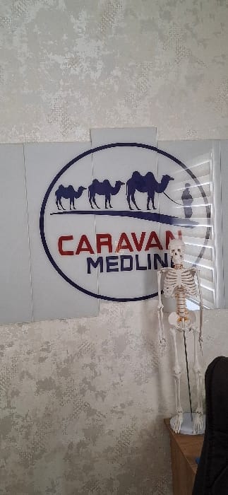 An interior scene in Urgench, Uzbekistan, features a light-toned wall covered with textured wallpaper. Centrally positioned is a multi-panel rectangular sign with a circular logo. The logo, outlined in dark blue on a white background, depicts the dark blue silhouettes of three camels of ascending size walking left to right, followed by a human figure holding a lead to the largest camel. Below the animal figures, the word "CARAVAN" is displayed in red, above "MEDLINI" in dark blue. To the immediate right of the sign, a life-sized, off-white anatomical human skeleton model stands on a dark circular base, exhibiting gold-colored joint articulation points. The base of the skeleton rests on a light-brown wooden surface. A dark office chair with a black backrest is partially visible on the far right. Horizontal white vertical blinds are visible through a window opening behind the right side of the sign and skeleton model.