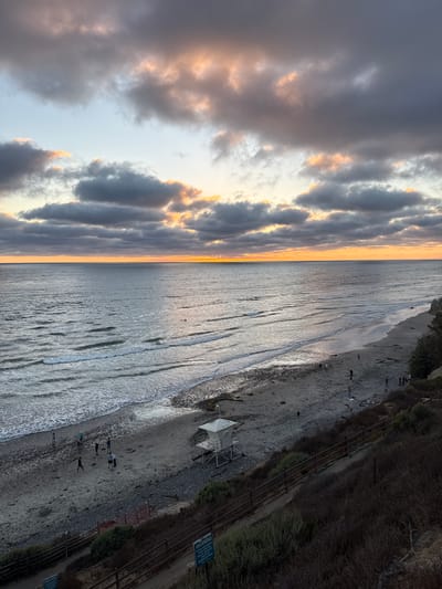 A wide-angle, elevated shot captures a beach scene at dusk in Encinitas, California. The sky is filled with dramatic, overcast clouds illuminated by the setting sun, which casts a golden glow along the horizon. The ocean stretches out, with gentle waves rolling onto the sandy and pebbly shore. Several individuals are present on the beach, some walking, others gathered in small groups. A white lifeguard tower stands on the sand near the water's edge. A winding pathway or stairs, bordered by vegetation and a wooden fence, leads down from an elevated vantage point, where the photograph was taken. The overall mood is serene and picturesque.