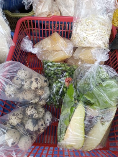 A red plastic basket contains various food items packaged in clear plastic bags. These include two bags filled with speckled quail eggs, a bag of small green peas, two bags of translucent yellow jelly-like squares, a bag of bean sprouts, and a bag containing green leafy vegetables with two pieces of light green fruit. The basket sits on a blue and white checkered surface. Additional bags of unidentified items are visible in the background.