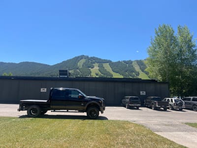 A black flatbed Ford truck is parked on a grassy area in front of a dark, corrugated metal building. Behind the building, a mountain is visible with ski slopes cut into its forested slopes. Several other trucks and SUVs are parked in a paved lot to the right of the Ford truck. The sky is clear and blue, indicating a sunny day. The scene appears to be set in a rural or mountainous area, likely during the daytime. There is a sign on the building with text, but it is too small to be legible. The overall mood is calm and clear.