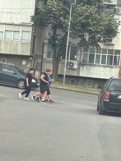 The image captures a street scene in the city of Aytos, Bulgaria. In the foreground, a gray asphalt road is visible, with a few small white debris pieces scattered across its surface. Three individuals are crossing the road from left to right. The person on the far left, wearing all black attire, walks with their right leg forward. Following closely behind are two other individuals, one in a blue shirt and black shorts, carrying a blue backpack, and another in a black t-shirt and black shorts. The person in the blue shirt is holding a small object in their right hand. To the left of the pedestrians, a dark blue car is partially visible, moving in the same direction. In the background, a multi-story building with a rough, gray facade dominates the scene. The building has several windows with horizontal blinds and air conditioning units mounted on its exterior. A large tree with green foliage partially obscures the building. To the right of the building, a black Volkswagen Passat station wagon is parked on the side of the road, facing away from the viewer, with its rear visible. The license plate is partially obscured. The sky is overcast.