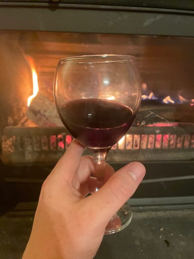 The image depicts a human hand holding a wine glass containing dark red liquid in front of an active fireplace, set in Palež, Montenegro. The hand, exhibiting light-toned skin, is positioned in the lower-center of the frame, grasping the stem of the clear glass. The glass holds an estimated 80-100 ml of liquid. The background is dominated by the interior of a fireplace or wood-burning stove, where orange and blue flames are visible consuming logs. Red-hot embers glow at the base of the fire, resting on a dark-colored grate. Reflections from the fire are visible on the surface of the wine and the glass itself. The scene is illuminated primarily by the firelight.