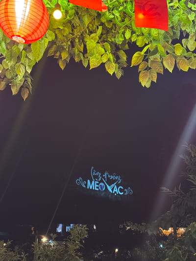 The scene captures an outdoor nighttime ambiance in the Mèo Vạc Commune, Vietnam. The left of the frame is filled with lush foliage, intertwined with a red communist flag and a traditional red lantern emitting a warm glow. The central part of the image transitions to a darker space, illuminated by a faint beam of light, which seems to project a light-blue illuminated script “Mèo Vạc”. On the right side, there is more foliage, and faint distant structures suggesting the presence of buildings.