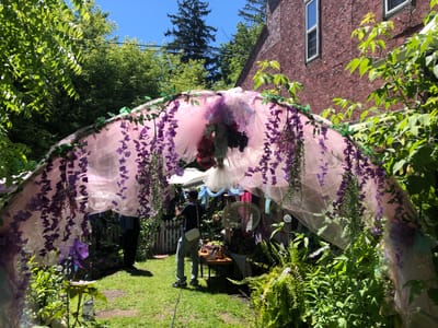 In the city of Severn, Canada, a person stands on a grassy area, holding a mobile device to take a photograph. The person is wearing a dark t-shirt, blue jeans, and white sneakers, with a white crossbody bag. They are positioned under a large, arching structure draped with sheer pink fabric and adorned with artificial purple flowers and greenery. The structure appears to be part of a backyard garden setup, with a white picket fence visible behind it. Various decorative elements are present within the arch, including a white fan, a pinwheel, and several artificial floral arrangements. The surrounding environment is lush with green trees and foliage, under a clear blue sky. A brick building with windows is visible to the right of the scene.