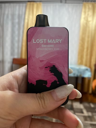 A person's hand with manicured nails is holding a pink vape device. The device has black lettering that reads "LOST MARY BM16000 STRAWBERRY KIWI". The background is blurred and shows curtains and furniture. There is no discernible indication of the location being in Зыбинское сельское поселение, Ukraine.