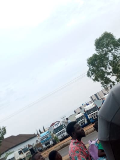 This is a ground-level, slightly tilted, outdoor photograph taken on a cloudy day in Wuro Dole, Nigeria. In the foreground, a man with dark skin and a beard is visible in profile, wearing a red and green floral print shirt. He is looking to his right, with his head slightly tilted upwards. To his right, a purple backpack is partially visible. Behind him, another person's head is visible.
In the mid-ground, several large trucks are parked along a road. These include white trucks with blue accents, and at least one white truck with a blue cab. Buildings and trees are visible in the background. One building appears to be a warehouse or industrial structure with a light-colored facade and dark stripes. Another building has a brown, sloped roof. The sky is overcast, with a hint of power lines visible in the upper portion of the image. The overall impression is of a commercial or transportation hub in an urban or semi-urban environment.