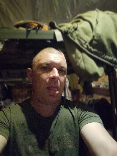 This is a medium shot, selfie-style photograph of a man with a shaved head, wearing a dark t-shirt underneath a green t-shirt. He is looking directly at the camera with a neutral expression, his tongue slightly extended. The background appears to be an interior space, possibly a military bunk or storage area, with green fabric or blankets visible. There are shelves with objects on them in the upper left of the frame, and more green fabric or equipment on the right. The lighting is dim, suggesting it might be nighttime or an indoor environment with limited light. The overall impression is of a candid and informal moment captured. There is no visible text in the image.