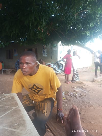 The image depicts an outdoor scene in what appears to be a rural or semi-urban area in Dokan Tofa, Nigeria. In the foreground, a man wearing a yellow t-shirt with a black logo and dark pants is seated. He is looking to his left. His left hand, adorned with a beaded bracelet, rests on a wooden surface. His feet are visible in the bottom right of the frame, positioned on another wooden surface.

In the middle ground, two women are standing near a motorcycle. One woman, on the left, wears a red t-shirt and a pink skirt. The other woman, to her right, is in a white t-shirt. Behind them, a building with visible posters on its exterior wall is present.

Further in the background, other individuals are visible, including a person in an orange shirt on the left and another person seated in the middle ground. The environment is dusty with scattered debris and vegetation. A large tree with dense foliage provides shade over the scene. The date and time "06/16/2026 17:16" are superimposed on the lower right corner of the image.